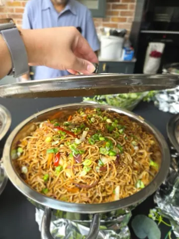 A chafing dish of hakka noodles with peppers and spring onion, the lid just lifted.