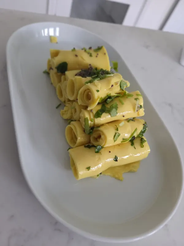 Rolled khandvi on a white plate, scattered with coriander and mustard seed.